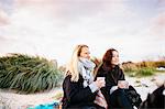 Friends looking away while holding coffee mugs on beach against sky