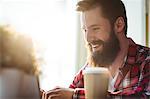 Cheerful young hipster with disposable cup and laptop at coffee house