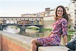 Remarkable holiday in Florence. Smiling young woman in a dress sitting on the embankment near Ponte Vecchio