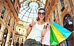 Discover most unexpected trends in Milan. Portrait of happy fashion woman in eyeglasses with colorful shopping bags in Galleria Vittorio Emanuele II showing thumbs up