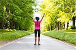 Beautiful mixed race African American young woman girl teenager fitness running jogging celebrating success on road lined with spring or summer green trees
