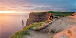 Cliffs of Moher (Aillte an Mhothair), Doolin, County Clare, Munster province, Ireland, Europe. Panoramic view with a woman watching the sun setting from the top of the cliff.