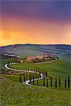 Valdorcia, Siena, Tuscany, Italy. Road of cypresses leading to a farmhouse with a stormy sunset in the background.