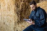 Farmer using digital tablet while sitting on hay bale at barn