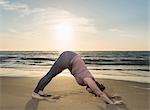 Mature woman practising yoga on a beach at sunset, downward facing dog pose