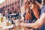 Young male hipster twins with red hair and beards reading smartphone texts at sidewalk bar