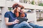 Young male hipster twins with red hair and beards sitting on bridge browsing digital tablet