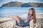 Beautiful young woman talking on smartphone reclining on armchair at beach, Costa Rei, Sardinia, Italy