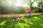 Mature woman mowing sunlit garden lawn with lawn mower