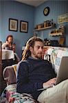 Hipster man using laptop while sitting on sofa with woman in background at home