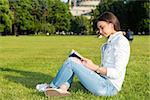 Smiling beautiful young woman on grass and reading book, against background of summer green park.