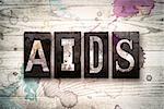 The word "AIDS" written in vintage dirty metal letterpress type on a whitewashed wooden background with ink and paint stains.