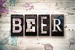 The word "BEER" written in vintage dirty metal letterpress type on a whitewashed wooden background with ink and paint stains.