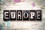 The word "EUROPE" written in vintage, dirty metal letterpress type on a whitewashed wooden background with ink and paint stains.