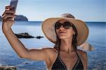 Young woman wearing sunhat on beach taking smartphone selfie, Villasimius, Sardinia, Italy