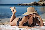 Young woman wearing sunhat chatting on smartphone whilst sunbathing on beach, Villasimius, Sardinia, Italy