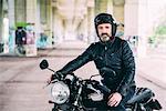 Mature male motorcyclist sitting on motorcycle wearing crash helmet under flyover