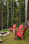Red plastic garden chairs around rock fire pit in backyard of cottage-style log home in summer, Quebec, Canada