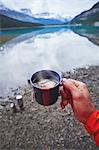 Cropped view of mans hand holding cup of tea by river, Banff, Alberta, Canada