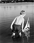 1960s YOUNG BOY WEARING BATHING SUIT STANDING IN WATER LAUNCHING TOY SAILING BOAT