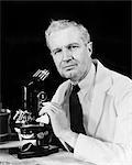 1940s MAN SCIENTIST USING MICROSCOPE WEARING A WHITE LAB COAT AND TIE LOOKING AT CAMERA