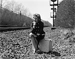 1950s 1960s WOMAN SAD WORRIED FACIAL EXPRESSION SITTING  ON SUITCASE BY SIDE RAILROAD TRACKS