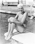 1930s GIRL WEARING BATHING SUIT AND SWIM CAP SITTING ON DIVING BOARD HOLDING KNEES LOOKING AT CAMERA