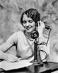 1920s SMILING WOMAN SITTING AT DESK WRITING AND TALKING ON CANDLESTICK TELEPHONE