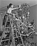 1950s JUVENILE GIRL ON LADDER TRYING TO PUT ANGEL ON TOP OF CHRISTMAS TREE