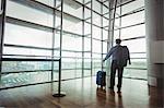 Rear view of businessman with luggage looking through glass window at airport