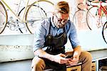 A man working in a bicycle repair shop sitting checking his phone for messages. Coffee break.