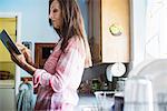 Senior woman with long brown hair standing in a kitchen, using a digital tablet.