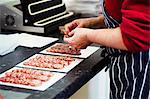 Close up of a butcher wearing a striped blue apron, packaging slices of salami.