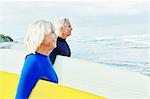 Senior woman senior man standing on a beach, wearing wetsuits and carrying surfboards.