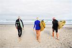Senior woman and two senior men on a beach, wearing wetsuits and carrying surfboards.