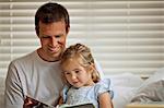 Father sitting on a bed reading a book with his daughter.