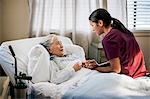 Friendly young nurse sits on an elderly patient's bed to chat with them.