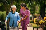 Friendly young nurse helps an elderly lady exercise with her walker in a sunny garden.