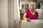 Anxious elderly woman gazes out of the kitchen window as she debates signing some paperwork.