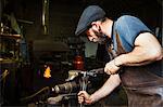 A blacksmith in a leather apron bends a cone of red hot metal in a vice using a wrench and a pair of tongs.