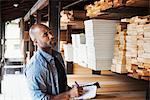 Man standing in a lumber yard, holding a folder, checking wood, writing notes.