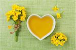 Rapeseed oil in heart shaped bowl and flowers on green background, top view.
