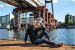 Pensive barefoot guy in glasses sits on the quay on the background of the river and the bridge. He wears blue jeans and a dark shirt. Male looks down. Outdoors. Horizontal.