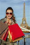 Portrait of cheerful young elegant woman in trench coat with shopping bags near Eiffel tower