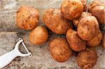 Pile of round baking potatoes beside peeler against a rustic background as seen from an overhead view