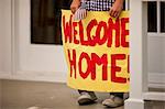 Welcoming sign held by a boy as he stands on the front porch of his home.