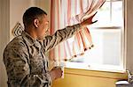 Smiling young adult soldier holding a mug while looking out the window of his home.