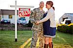 Smiling soldier standing with his pregnant wife next to a real estate sign in the front yard of their home.