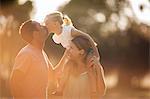 Young girl sitting on her mother's shoulders leaning over to kiss her father.