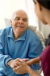 Senior man holding hands with a female nurse while sitting in an arm chair.